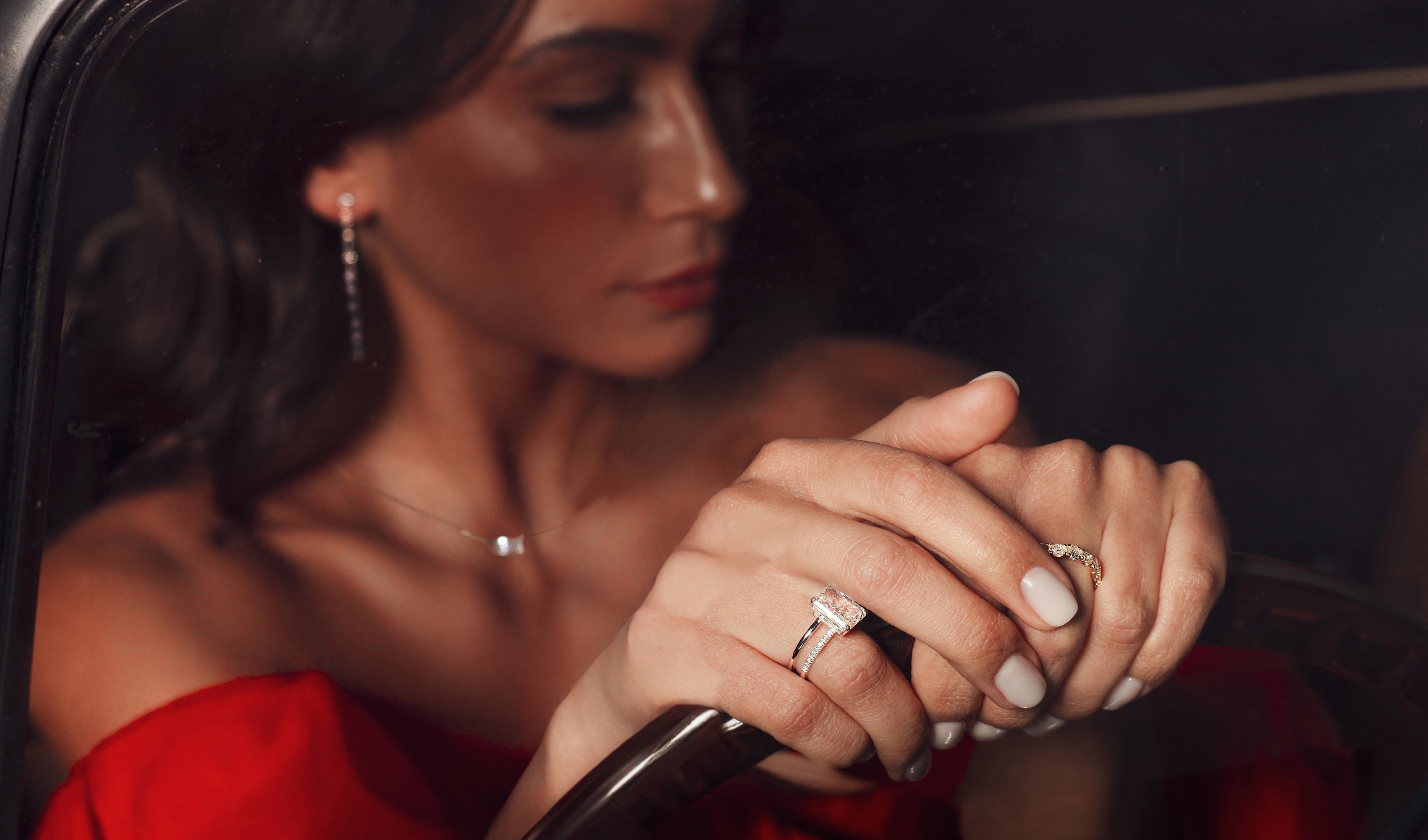 Eine Frau in einem roten Kleid hält ein Lenkrad. Ihre Nägel sind weiß lackiert und sie trägt einen Diamantring und einen kleinen Ring. Ihre Ohrringe und ihre Halskette sind ebenfalls sichtbar. Die Beleuchtung ist warm und hebt ihr Profil sanft hervor.