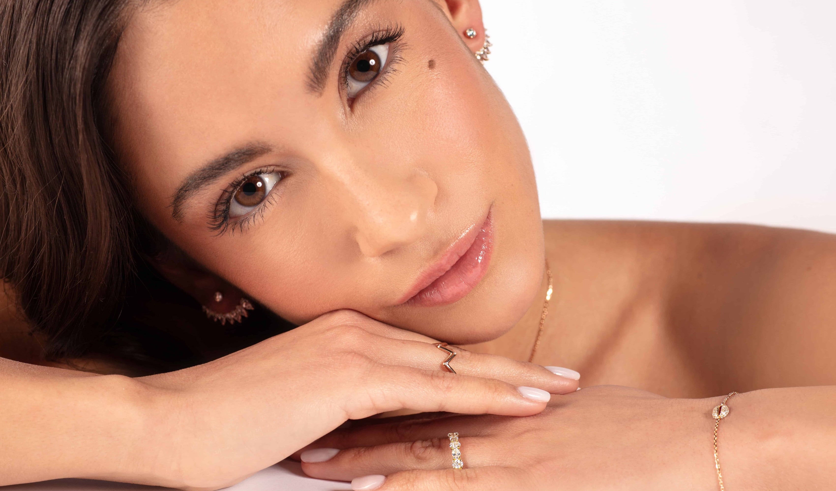 Eine Frau mit braunem Haar stützt sich auf ihre Hände und präsentiert filigrane Ohrringe, Ringe und das Atelier Sofia RHEA ROSÉGOLD Armband, das mit im Labor gezüchteten Brillanten verziert ist. Ihr ruhiger Gesichtsausdruck und ihr natürliches Make-up heben sich vor einem hellen Hintergrund ab.