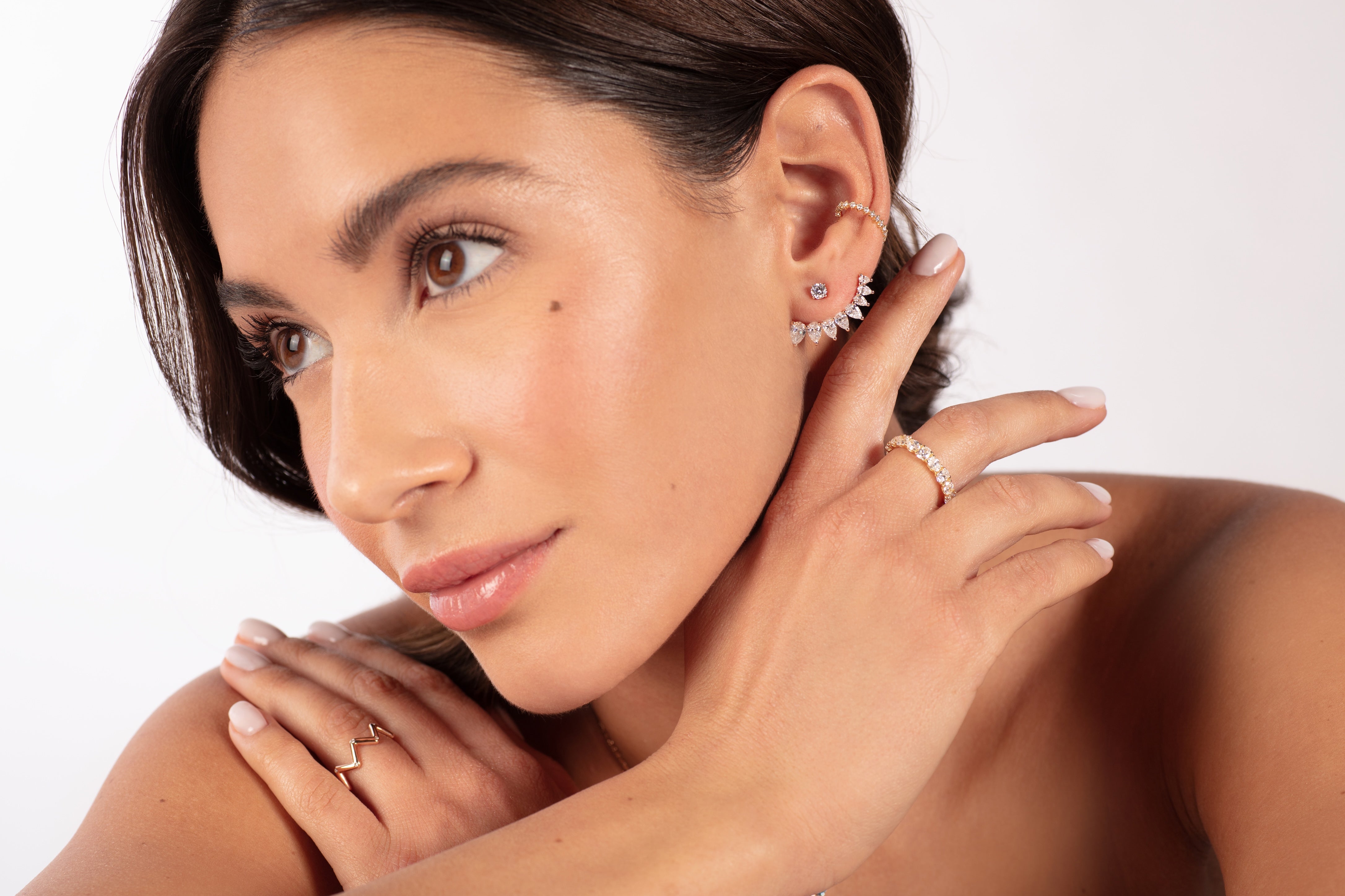 Eine braunhaarige Frau berührt ihren Hals und ihre Schulter und präsentiert mehrere Ohrringe und einen atemberaubenden KALLIOPI WEIßGOLD-Ring von Atelier Sofia. Ihr dezentes Make-up hebt ihre Augen und ihren glatten Teint vor einem weißen Hintergrund hervor und betont die im Labor gezüchteten Diamanten und ihren heiteren Ausdruck.