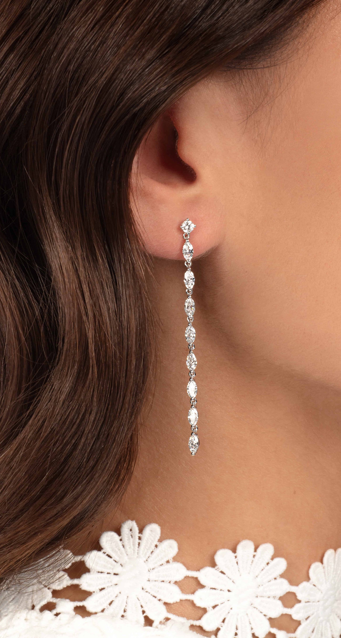 Nahaufnahme des Ohrs einer Frau, die einen langen, zarten Diamantohrring mit mehreren Steinen trägt. Das braune Haar der Frau ist hinter das Ohr gesteckt und sie trägt ein weißes Spitzentop mit floralen Details.