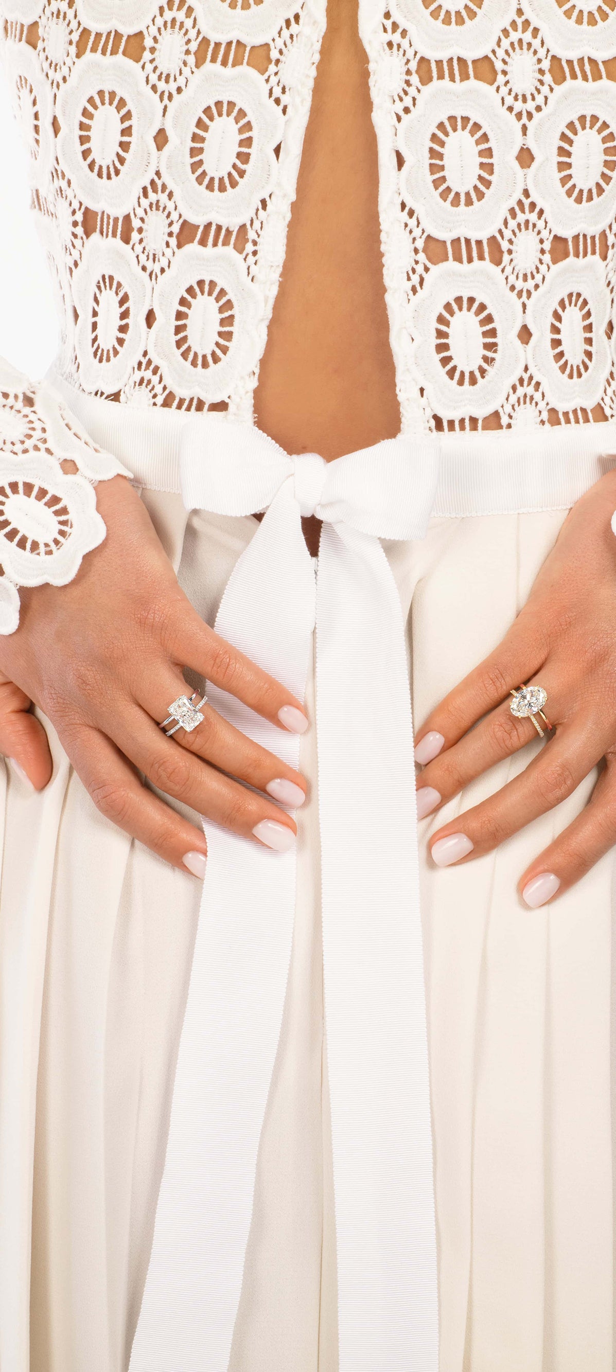 Eine Person trägt ein weißes, langärmliges Spitzenoberteil mit tiefem Ausschnitt, das mit einem Band zusammengebunden ist, und einen fließenden Rock. Ihre Fingernägel sind manikürt und sie trägt zwei Ringe mit großen Steinen an den Fingern.