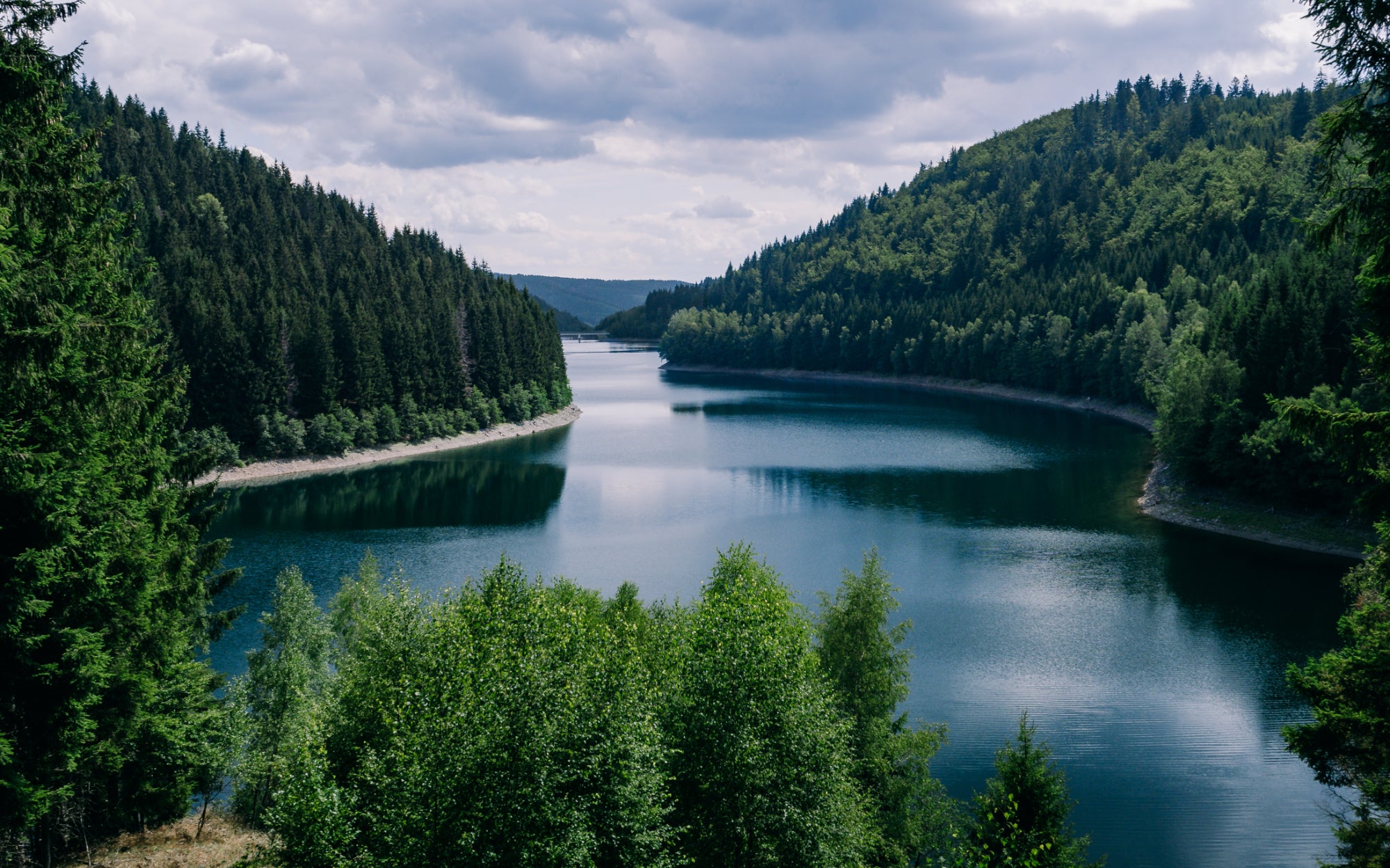 Ein ruhiger Fluss schlängelt sich unter einem bewölkten Himmel zwischen bewaldeten Hügeln hindurch. Das üppige Grün umgibt das ruhige blaue Wasser und schafft eine friedliche Naturlandschaft.
