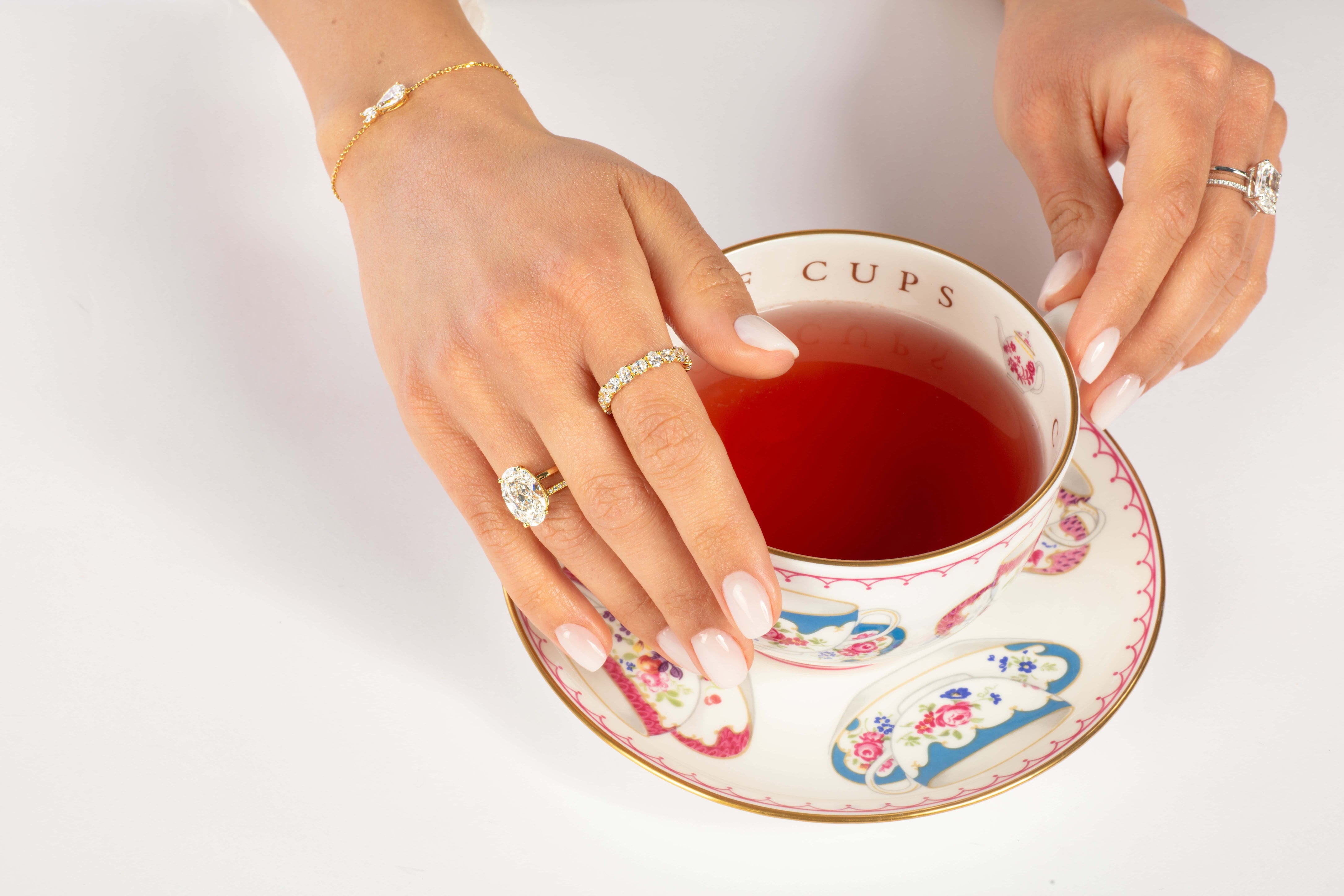 Hände mit eleganten Ringen und einem Armband halten eine mit Blumenmuster verzierte Teetasse, die mit einer roten Flüssigkeit gefüllt ist. Die Teetasse steht auf einer passenden Untertasse, beide sind mit farbenfrohen Mustern verziert. Der Hintergrund ist eine saubere weiße Oberfläche.