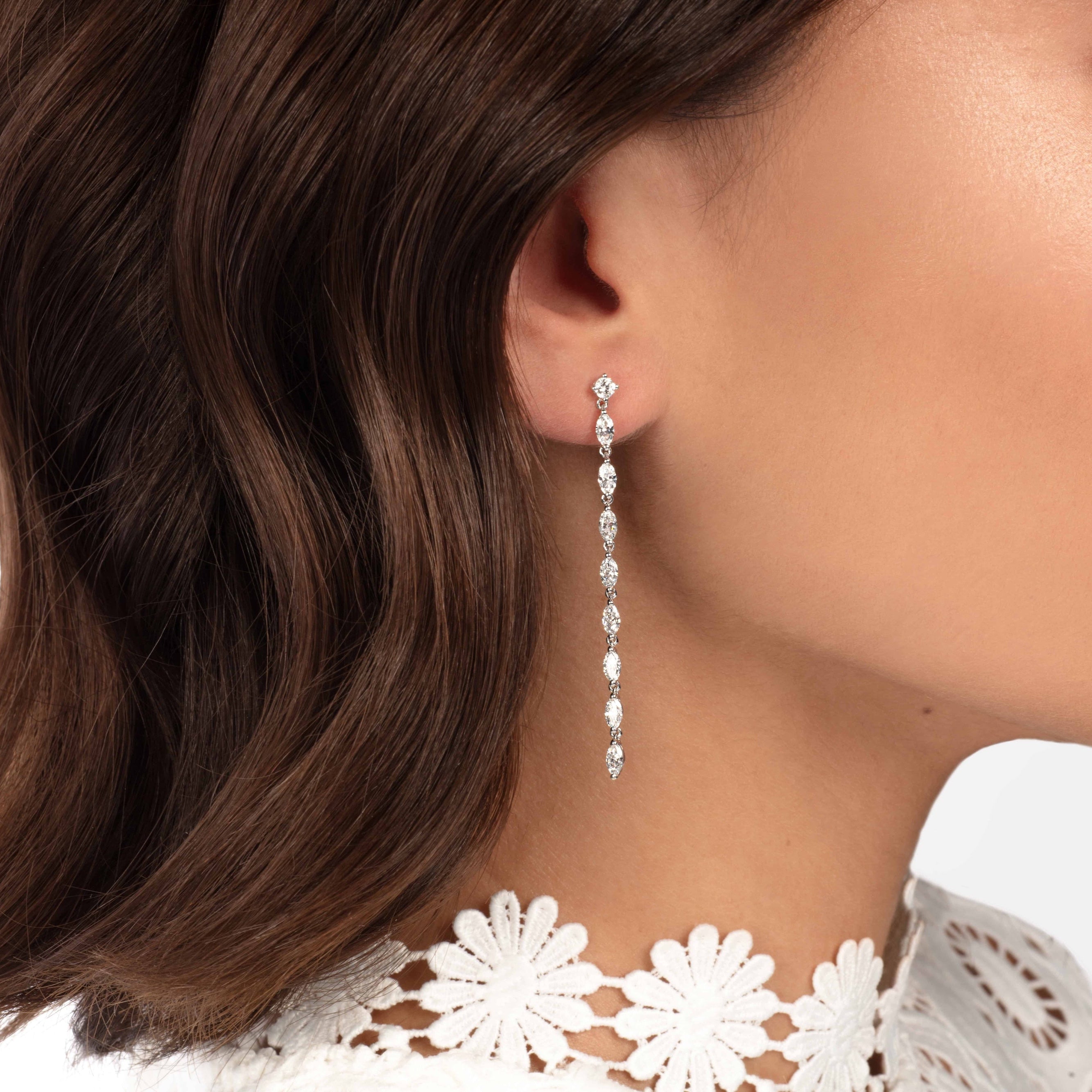 Nahaufnahme einer Frau, die Atelier Sofias AGLAIA WEIßGOLD trägt, elegante lange, im Labor gezüchtete Diamantohrringe. Ihr braunes Haar fällt sanft herab und ergänzt ihr weißes Spitzenhalsband mit floralen Details. Der makellose Hintergrund verstärkt die Brillanz ihrer Accessoires und lässt sie hell strahlen.