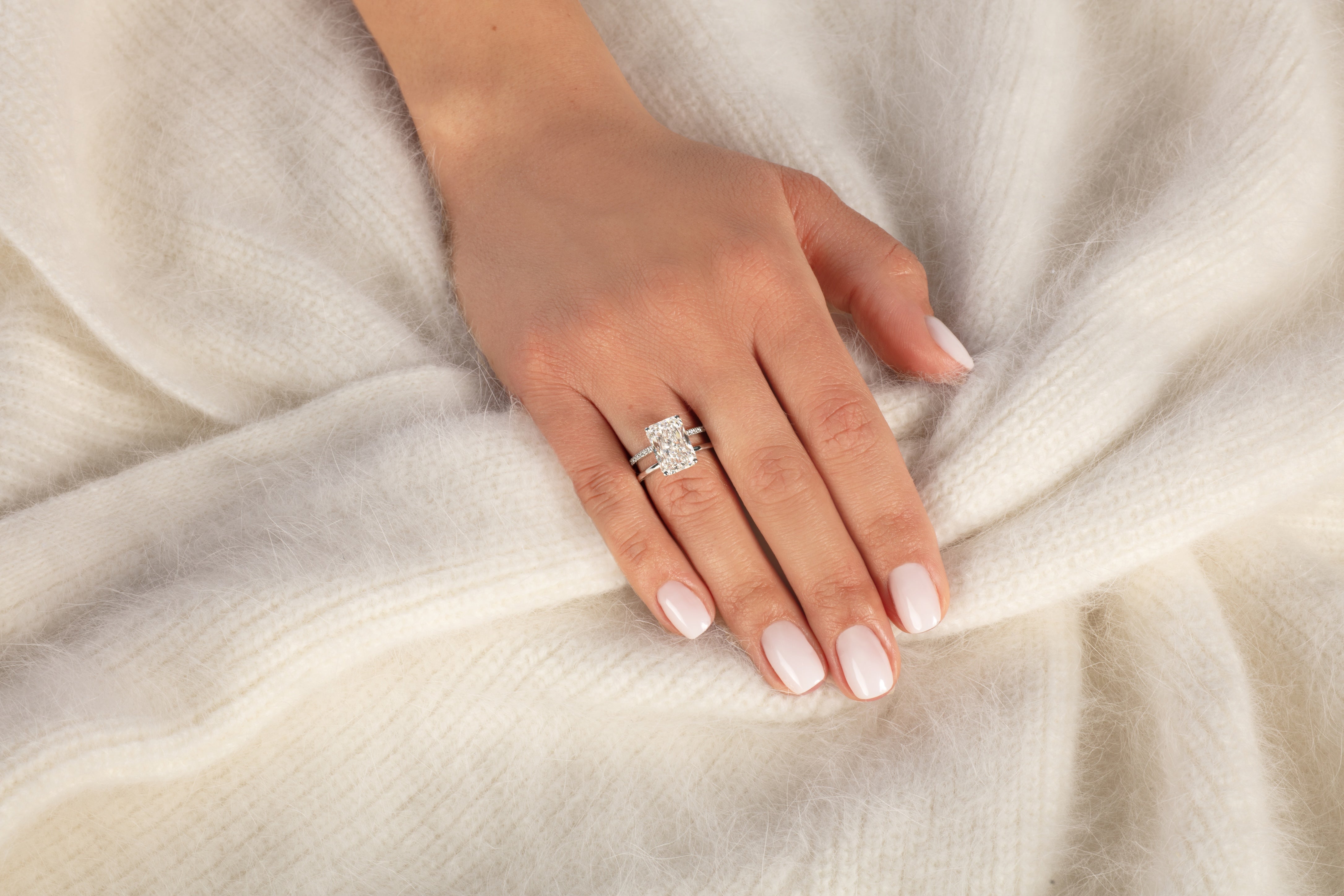 Eine manikürte Hand ruht auf einem weichen, weißen Stoff, geschmückt mit dem Ring und Ehering APHRODITE WEIßGOLD von Atelier Sofia, aufwendig gestaltet in 18 Karat Weißgold und funkelnd mit strahlend geschliffenen, im Labor gezüchteten Diamanten.