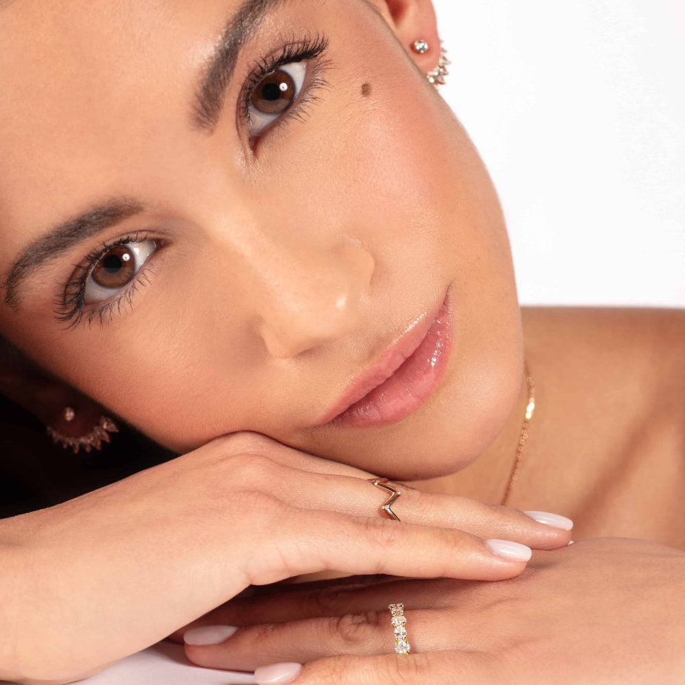 Eine Frau mit heller Haut stützt sich auf ihre Hand und lächelt sanft, während sie den Ring ATHENE ROSÉGOLD von Atelier Sofia und einen Zickzack-Ring trägt. Ihre braunen Augen und ihr dezentes Make-up heben sich vom schlichten weißen Hintergrund ab und zeigen ihre Eleganz.
